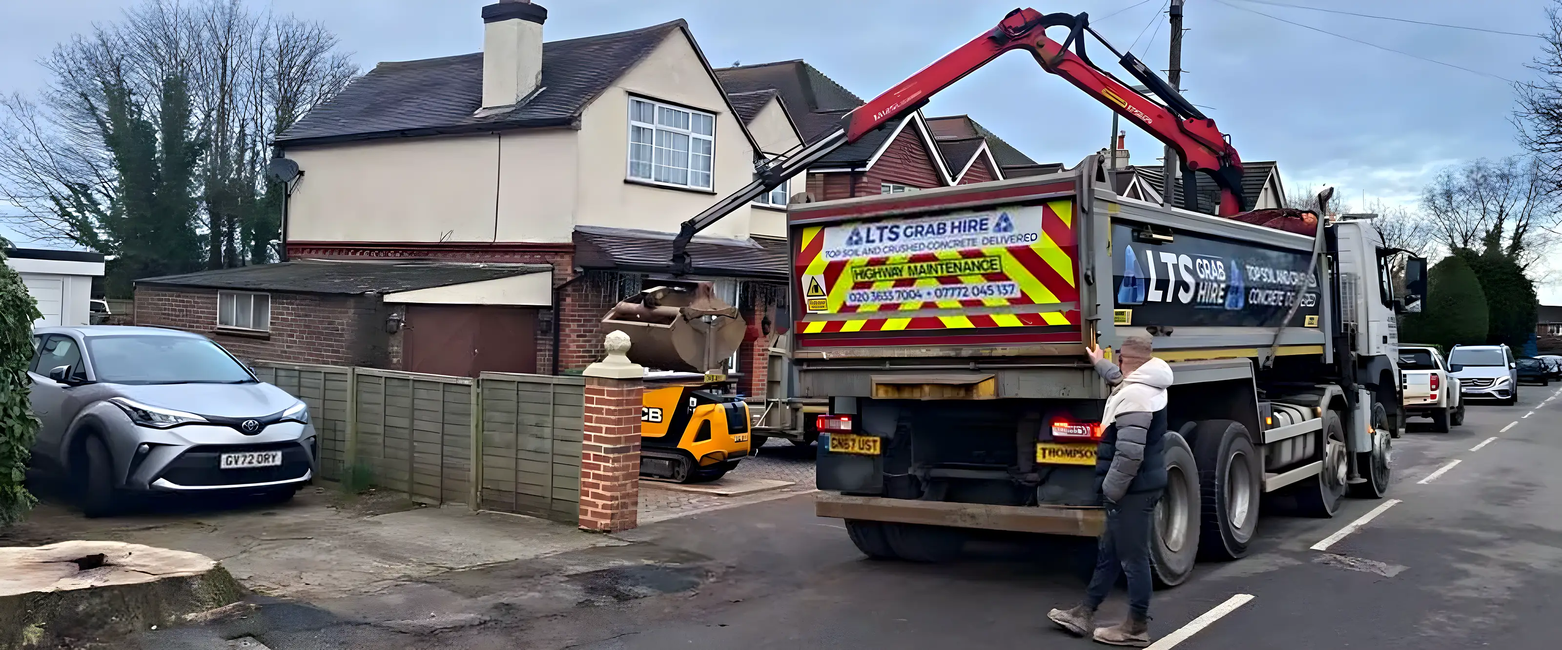 A grab lorry outside a house with the arm reaching over the wall to lift soil